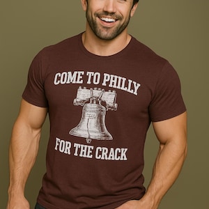 Może przedstawiać: Bordowa koszulka z napisem "COME TO PHILLY FOR THE CRACK" i grafiką Dzwonu Wolności. Osoba ma na sobie ciemne dżinsy i uśmiecha się. Tło jest jednolite oliwkowo-zielone.