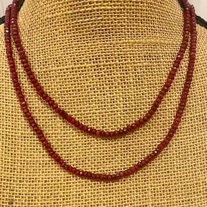May include: Two strands of a deep red beaded necklace. The beads are small and faceted, creating a sparkling effect. The necklace is displayed on a burlap-textured surface.