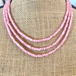 Puede incluir: Un collar de varias hebras con pequeñas cuentas redondas de color rosa claro. El collar se exhibe en un maniquí con un fondo de arpillera. Las cuentas están espaciadas uniformemente, creando un accesorio delicado y femenino.