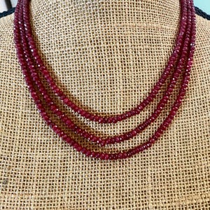 May include: A triple-strand necklace featuring small, deep red faceted beads. The necklace is arranged in three concentric layers, with each strand composed of numerous tiny, reflective, dark crimson stones that create a uniform, textured appearance.