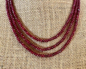 May include: A triple-strand necklace featuring small, deep red faceted beads. The necklace is arranged in three concentric layers, with each strand composed of numerous tiny, reflective, dark crimson stones that create a uniform, textured appearance.
