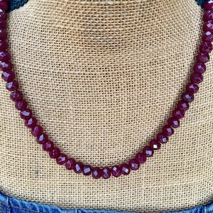 May include: A burgundy beaded necklace with faceted beads. The necklace is displayed on a burlap surface, with a blue denim garment visible at the bottom of the frame. The beads have a geometric shape and a glossy finish.