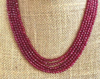 May include: A multi-strand necklace composed of small, faceted, deep red beads. The necklace is draped on a burlap-textured surface, showcasing the rich color and texture of the beads. The necklace is a statement piece.