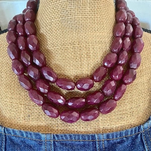 Op de afbeelding: Een ketting met drie strengen met grote, facetgeslepen, ovale kralen in een diepe bordeauxrode kleur. De ketting wordt getoond op een met jute bedekte buste, met een spijkergoed kledingstuk zichtbaar aan de onderkant.