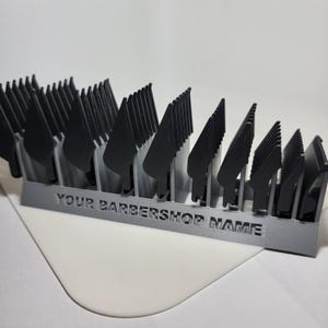Może przedstawiać: Szary organizer na ostrza do maszynek do strzyżenia z wieloma czarnymi ostrzami. Na froncie organizera wydrukowano napis "YOUR BARBERSHOP NAME". Organizer znajduje się na białej powierzchni.