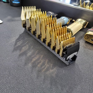 Könnte beinhalten: Ein Satz goldfarbener Haarschneidemaschinen-Klingenschutzvorrichtungen, in einer Reihe angeordnet, mit schwarzer Basis. Die Klingen sind abgewinkelt und haben ein scharfes, spitzes Design. Weitere goldene und schwarze Haarschneidemaschinen befinden sich im Hintergrund.