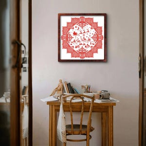 May include: A framed red and white Christmas-themed cross-stitch. The design features a detailed winter scene with a sleigh, reindeer, houses, and decorative borders. The artwork is displayed on a white wall above a wooden desk.