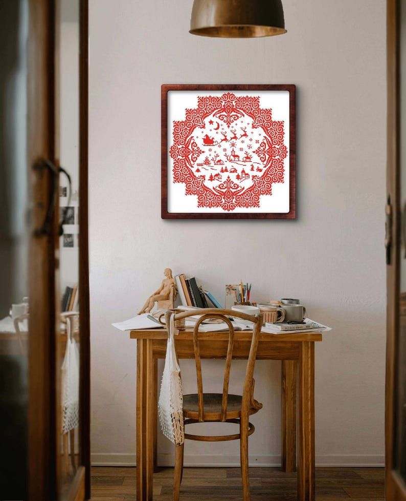 May include: A framed red and white Christmas-themed cross-stitch. The design features a detailed winter scene with a sleigh, reindeer, houses, and decorative borders. The artwork is displayed on a white wall above a wooden desk.