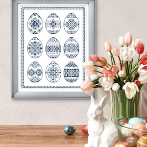 Op de afbeelding: Een ingelijste print met negen blauw-witte paasei-ontwerpen. De eieren tonen verschillende sneeuwvlok- en bloempatronen. Een vaas met roze en witte tulpen, een wit konijnbeeldje en decoratieve eieren staan op een houten oppervlak.