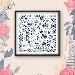 以下が含まれることがあります： 黒いフレームに入ったクロスステッチのサンプル。ネイビーブルーのデザインには、花や動物のモチーフ、アルファベット、そして「Spring」の文字が含まれています。背景は、花のアクセントが付いた明るいベージュ色です。