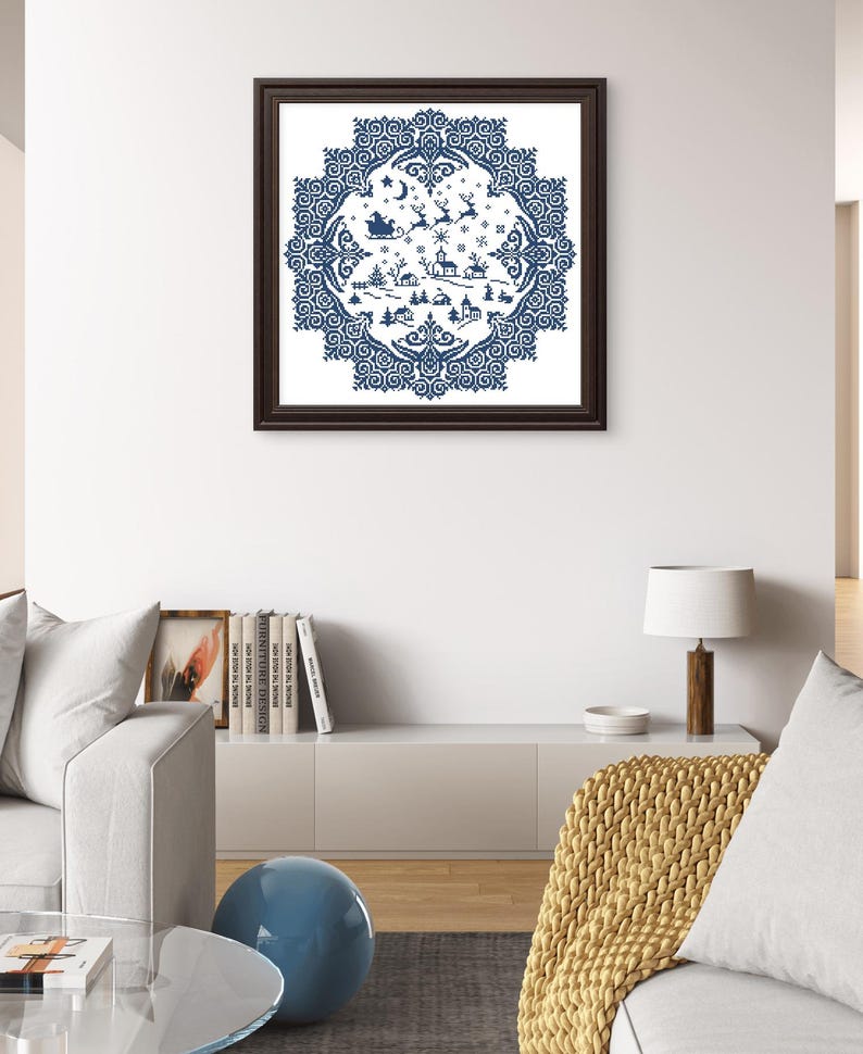 May include: A framed, blue and white Christmas-themed cross-stitch design. The artwork features a circular pattern with a winter village scene, reindeer, and Santa's sleigh. The frame is dark brown, and the artwork is displayed on a white wall.
