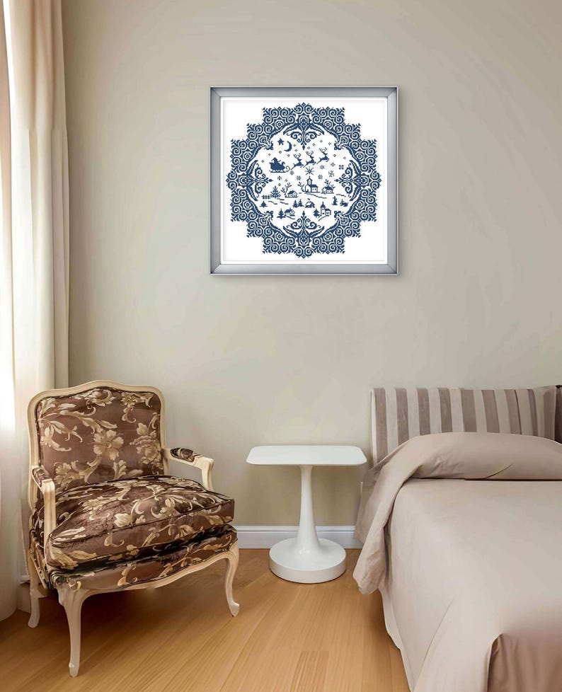 May include: A framed, blue and white cross-stitch artwork depicting a winter scene with Santa's sleigh, reindeer, and a village. The artwork is in a silver frame and hangs on a beige wall. A patterned chair and a white side table are in the foreground.