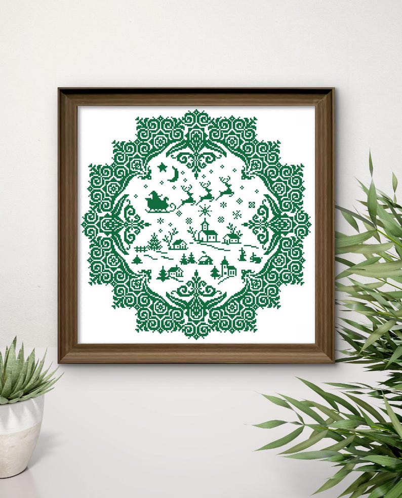 May include: A framed cross-stitch depicting a winter village scene in green thread on a white background. The design features a circular border with snowflakes, a sleigh, reindeer, houses, and trees. The frame is a dark brown color.