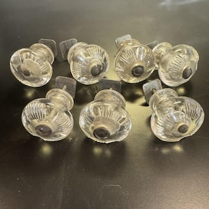 May include: Eight clear glass cabinet knobs with a ribbed design and dark centers. Each knob has a metal mounting plate attached. The knobs are arranged on a dark surface.