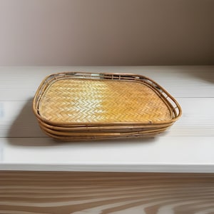 May include: A rectangular woven bamboo tray with rounded corners. The tray has a light brown color and a woven pattern. The tray is sitting on a white surface.