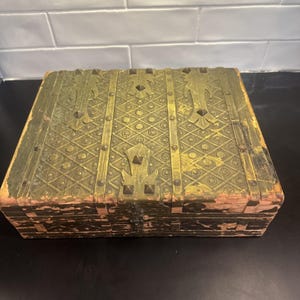 May include: A rectangular wooden box featuring a gold-toned metal overlay on the lid with a repeating diamond and floral pattern. The metal is secured with small studs and decorative accents. The front panel shows dark wood with a central metal latch.