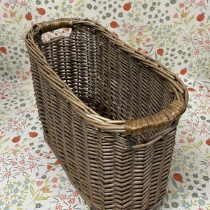 May include: A tall, woven wicker basket with a rectangular base and rounded top. The basket is a mix of light and dark brown tones, with a handle on each side. The basket is empty and sits on a floral patterned surface.