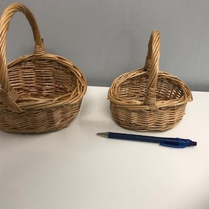 May include: Two woven wicker baskets with handles. The larger basket is oval shaped and the smaller basket is round. Both baskets are made of natural brown wicker.