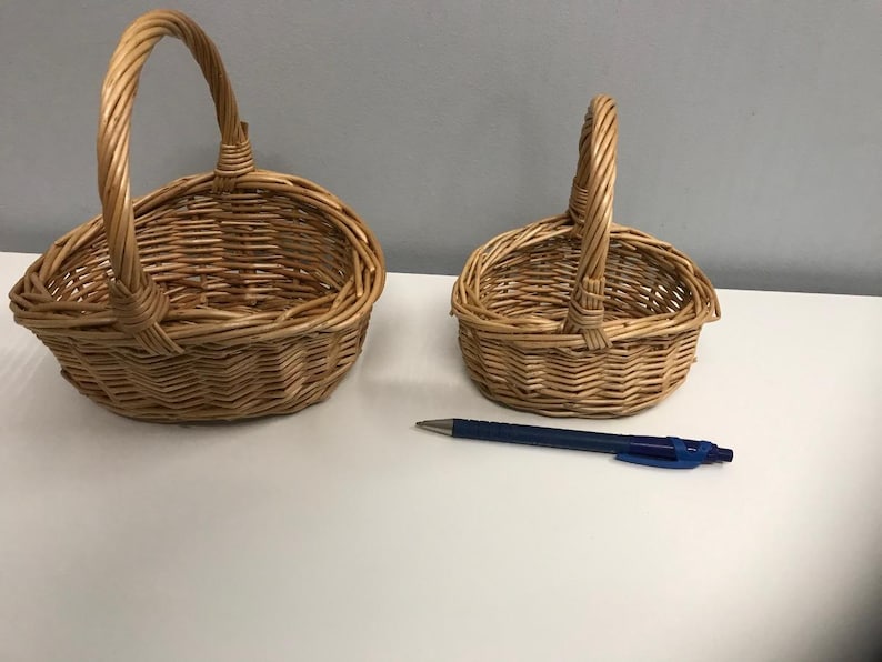 May include: Two woven wicker baskets with handles. The larger basket is oval shaped and the smaller basket is round. Both baskets are made of natural brown wicker.
