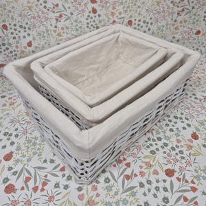 May include: Three white wicker storage baskets of different sizes, nested together. Each basket has a removable, off-white linen liner. The baskets are set on a floral patterned surface.