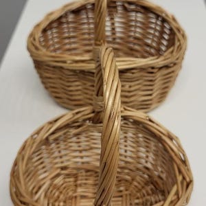 May include: Two oval-shaped wicker baskets with handles. The baskets are stacked on top of each other. The top basket is slightly larger than the bottom basket.