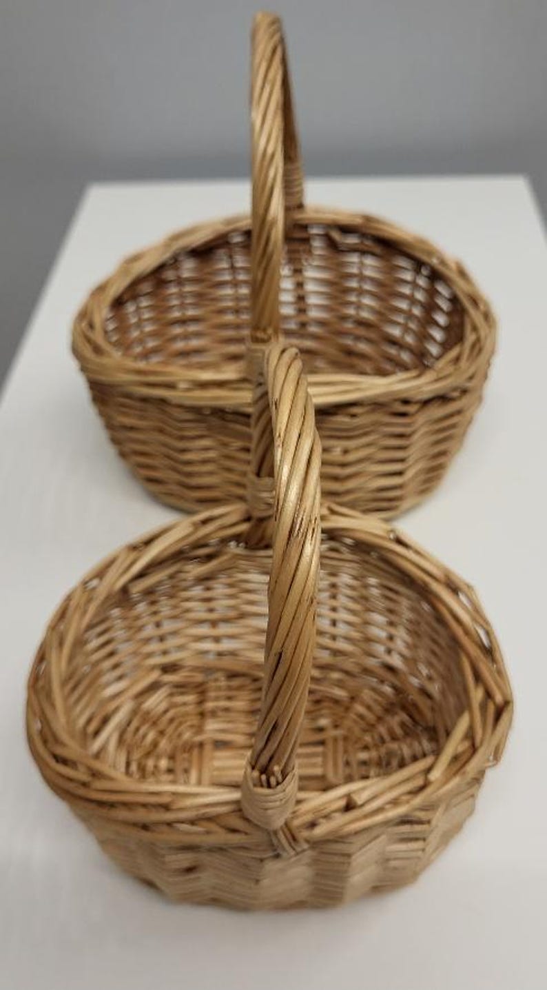 May include: Two oval-shaped wicker baskets with handles. The baskets are stacked on top of each other. The top basket is slightly larger than the bottom basket.