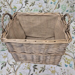 May include: A rectangular wicker basket with woven sides and two curved handles. The interior is lined with a tan burlap fabric. The basket is empty and sits on a floral patterned surface.