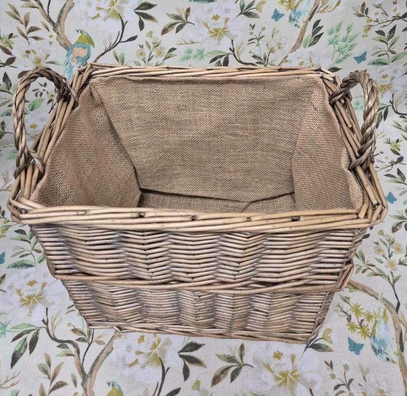 May include: A rectangular wicker basket with woven sides and two curved handles. The interior is lined with a tan burlap fabric. The basket is empty and sits on a floral patterned surface.