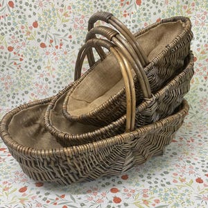 May include: A set of three nested oval baskets made of dark brown woven wicker, each featuring a sturdy arched handle and a fitted tan burlap fabric liner. The baskets are stacked together, showcasing a consistent chevron-like weave pattern.