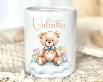 Könnte beinhalten: Weiße Keramik-Spardose mit einer Aquarell-Illustration eines Teddybären mit blauer Schleife, der auf einer Wolke sitzt. Der Name "Valentin" steht auf der Spardose. Der Bär ist von Alphabetblöcken umgeben.