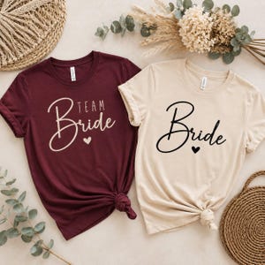 Könnte beinhalten: Ein kastanienbraunes T-Shirt mit dem Text Team Bride und einem kleinen Herz in Hellrosa sowie ein beiges T-Shirt mit dem Text Bride und einem kleinen Herz in Schwarz. Beide T-Shirts haben kurze Ärmel, einen Rundhalsausschnitt und einen Knoten am Saum.