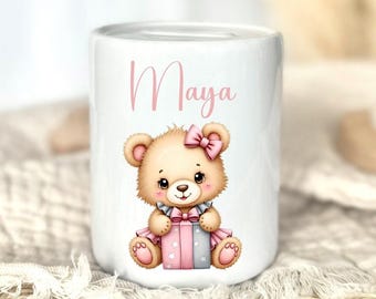 Könnte beinhalten: Weiße Keramik-Spardose mit einer Illustration eines Teddybären mit rosa Schleife und Kleid. Der Name "Maya" steht in rosa Schrift über dem Bären. Die Spardose ist zylindrisch und hat einen Schlitz für Münzen.
