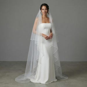 May include: A bride in a white strapless wedding dress and a long, sheer veil. The veil has a delicate trim and falls to the floor. The dress is form-fitting and simple, with a slight train. The background is a neutral gray.