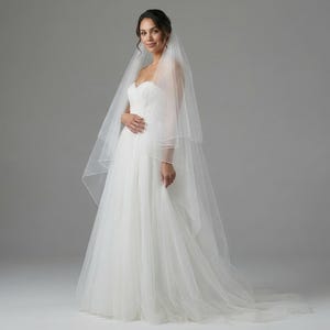 May include: A woman in a white wedding dress and veil poses in a studio. The strapless gown has a fitted bodice and a flowing skirt. The veil is sheer and drapes over the shoulders. The background is a plain gray.