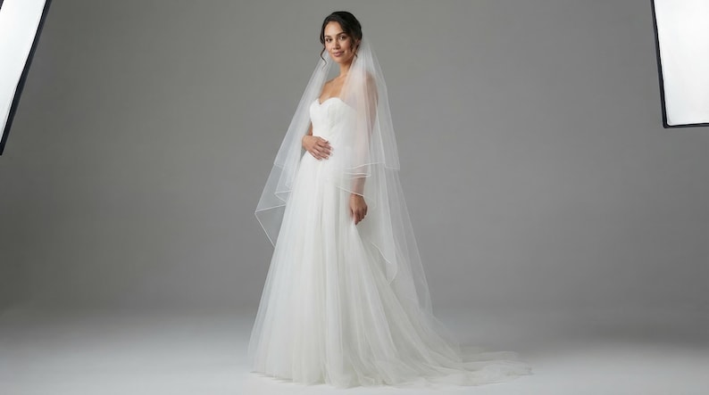 May include: A woman in a white wedding dress and veil poses in a studio. The strapless gown has a fitted bodice and a flowing skirt. The veil is sheer and drapes over the shoulders. The background is a plain gray.