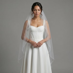 May include: A white wedding dress with a sweetheart neckline and a flowing skirt. The dress is paired with a sheer, white veil. The bride is standing in front of a gray background.