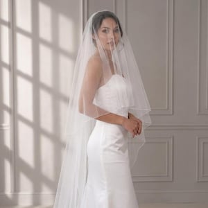 May include: A two-tier white bridal veil made of sheer, lightweight tulle fabric. The veil features a delicate, finished edge and drapes over the head and shoulders, extending down the back in a soft, flowing silhouette over a white dress.