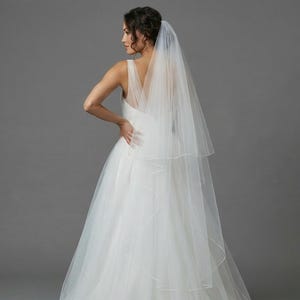 May include: A white wedding dress with a layered tulle skirt and a V-neck bodice. A sheer, two-tiered veil is draped over the shoulders. The bride is turned away from the camera, with her hand on her hip. The background is a solid gray.