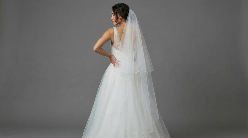 May include: A white wedding dress with a layered tulle skirt and a V-neck bodice. A sheer, two-tiered veil is draped over the shoulders. The bride is turned away from the camera, with her hand on her hip. The background is a solid gray.