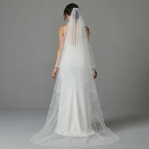 May include: A bride in a white wedding dress and veil, viewed from the back. The veil is a two-tiered design, with a delicate, sheer fabric. The dress is a simple, elegant style. The background is a plain, light gray.
