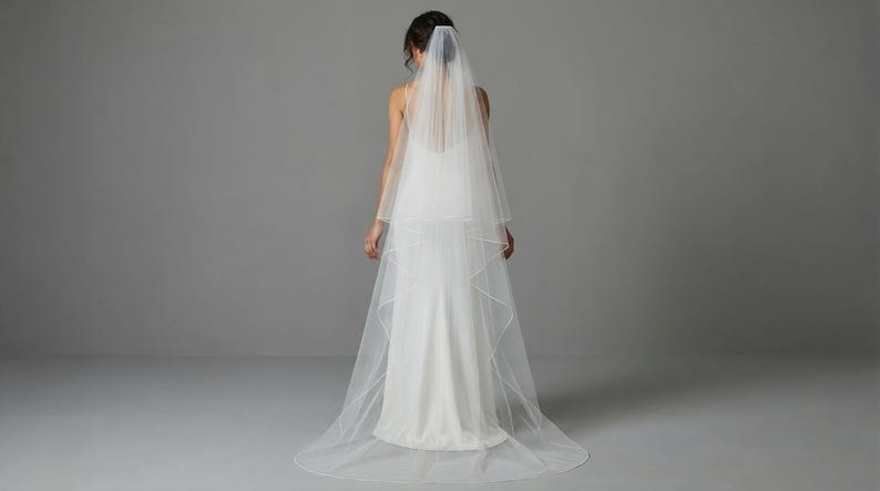 May include: A bride in a white wedding dress and veil, viewed from the back. The veil is a two-tiered design, with a delicate, sheer fabric. The dress is a simple, elegant style. The background is a plain, light gray.