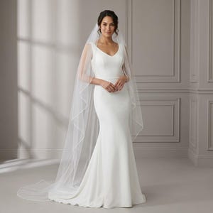 May include: A floor-length white wedding dress with a V-neckline and a fitted bodice that flares into a trumpet skirt. The dress is paired with a long, sheer white tulle veil that features a delicate finished edge, draping over the shoulders.