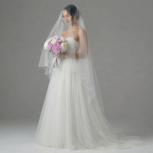 May include: A bride in a strapless white wedding gown, holding a bouquet of pink and white flowers. She is wearing a sheer veil. The image is taken in a studio setting with soft lighting.