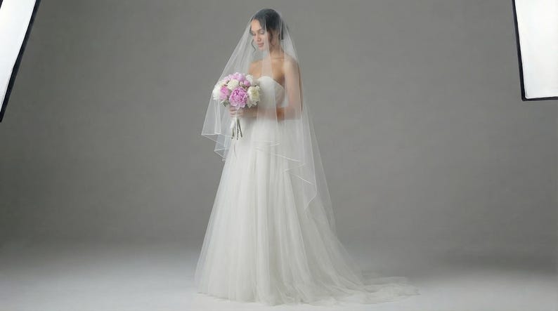 May include: A bride in a strapless white wedding gown, holding a bouquet of pink and white flowers. She is wearing a sheer veil. The image is taken in a studio setting with soft lighting.