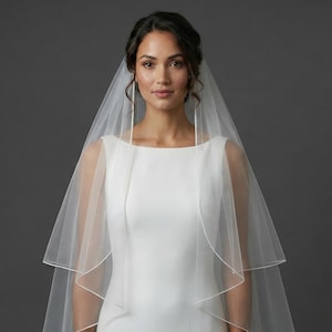 May include: A bride wearing a white wedding dress with a square neckline and a sheer, layered veil. The veil drapes over the shoulders and frames the face. The background is a solid gray.