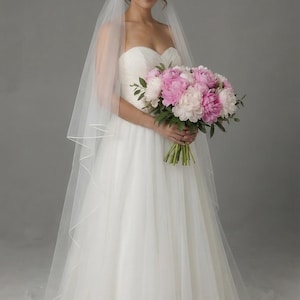 May include: A bride in a white strapless wedding dress and veil holds a bouquet of pink and white peonies. The dress has a flowing skirt. The background is a solid gray.
