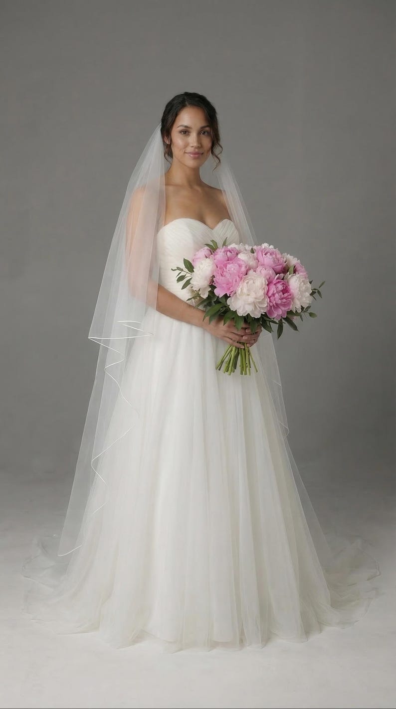 May include: A bride in a white strapless wedding dress and veil holds a bouquet of pink and white peonies. The dress has a flowing skirt. The background is a solid gray.