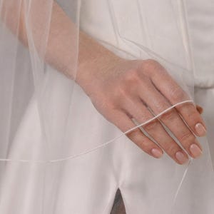 May include: A sheer, white bridal veil with multiple layers. The veil is held by a hand, showcasing the delicate fabric and design. The bride's white dress is visible in the background.