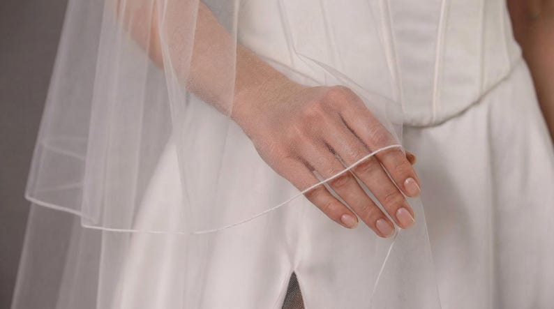May include: A sheer, white bridal veil with multiple layers. The veil is held by a hand, showcasing the delicate fabric and design. The bride's white dress is visible in the background.