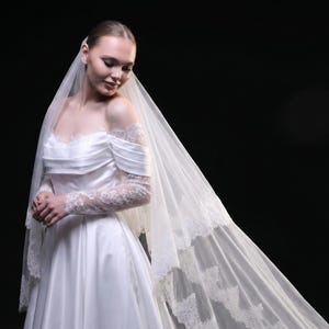 Op de afbeelding: Een bruid in een witte satijnen trouwjurk met een off-the-shoulder halslijn en lange kanten mouwen. Een lange, vloeiende sluier met kanten rand valt naar beneden. De achtergrond is effen zwart.